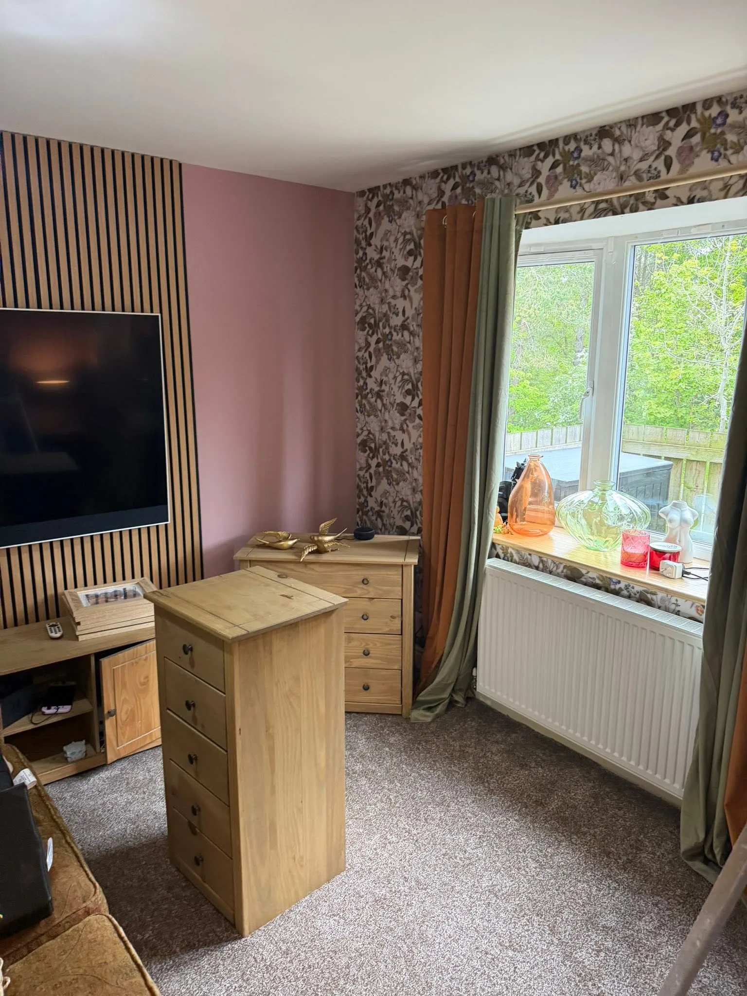 Room with an oak slat wall behind a wall-mounted TV, a dusky pink wall and floral wallpaper
